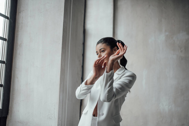 Young adult beauty swarthy woman in formal evening white suit no shirt hand on face touching. Stylish black curly hair sensual african american model fashionista posing at studio in fashion pantsuit - Фото, зображення