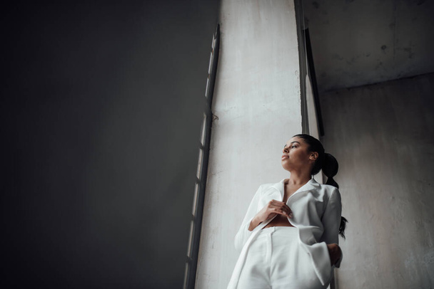 Young adult beauty swarthy woman in formal evening white suit with bra at thoughtful. Stylish black curly hair sensual african american model fashionista posing at studio in fashion pantsuit - 写真・画像