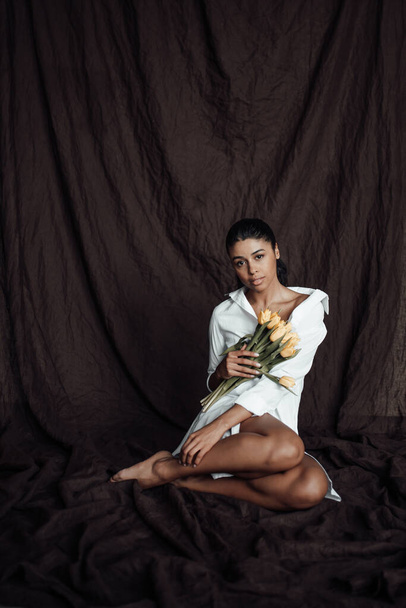 Young adult beauty swarthy woman in white shirt bare shoulder hugs bouquet of yellow tulips. Stylish black curly hair sensual african american model fashionista posing in studio at spring holidays - Fotografie, Obrázek