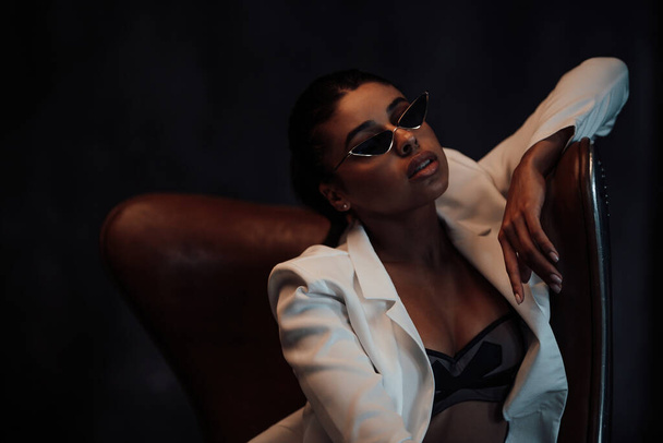 Young adult beauty swarthy woman in formal evening white suit with bra at cat eye sunglasses. Stylish black curly hair sensual african american model fashionista posing at studio in fashion pantsuit - Fotó, kép