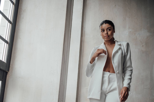 Young adult beauty swarthy woman in formal evening white suit with bra at thoughtful. Stylish black curly hair sensual african american model fashionista posing at studio in fashion pantsuit - Foto, afbeelding