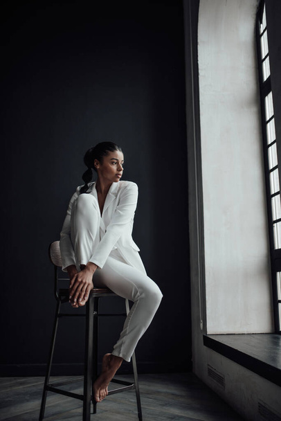 Young adult beauty swarthy woman in formal evening white suit with bra at thoughtful. Stylish black curly hair sensual african american model fashionista posing at studio in fashion pantsuit - Valokuva, kuva
