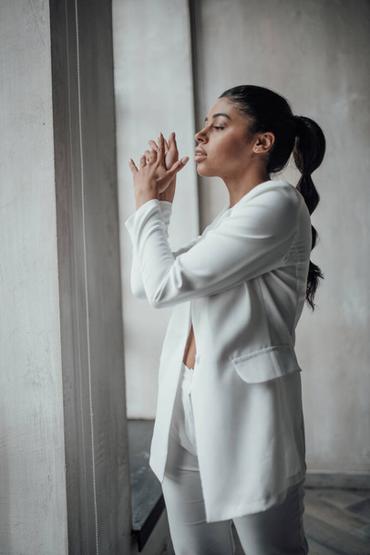 Young adult beauty swarthy woman in formal evening white suit no shirt hand on face touching. Stylish black curly hair sensual african american model fashionista posing at studio in fashion pantsuit - Foto, Imagem
