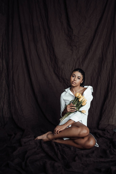 Young adult beauty swarthy woman in white shirt bare shoulder hugs bouquet of yellow tulips. Stylish black curly hair sensual african american model fashionista posing in studio at spring holidays - Foto, Bild