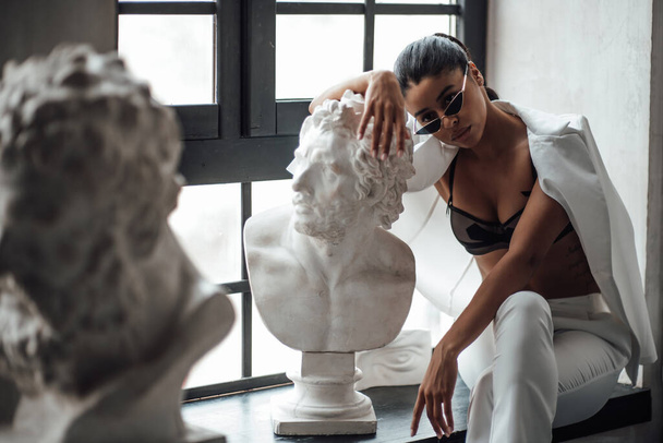 Young beauty swarthy woman in formal evening white suit no shirt hugs greek man bust statue. Stylish black curly hair sensual african american model fashionista posing at studio in fashion pantsuit - Photo, Image