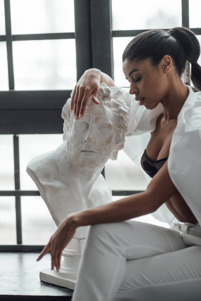 Young beauty swarthy woman in formal evening white suit no shirt hugs greek man bust statue. Stylish black curly hair sensual african american model fashionista posing at studio in fashion pantsuit - Foto, Imagem
