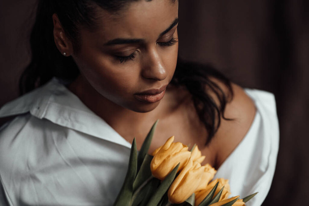 Giovane donna adulta bellezza swarthy in camicia bianca spalla nuda abbracci bouquet di tulipani gialli. Elegante capelli ricci neri sensuale modello afro americano fashionista in posa in studio alle vacanze di primavera - Foto, immagini
