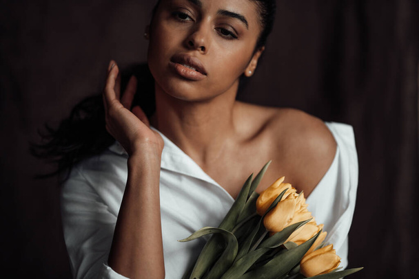 Young adult beauty swarthy woman in white shirt bare shoulder hugs bouquet of yellow tulips. Stylish black curly hair sensual african american model fashionista posing in studio at spring holidays - Photo, Image