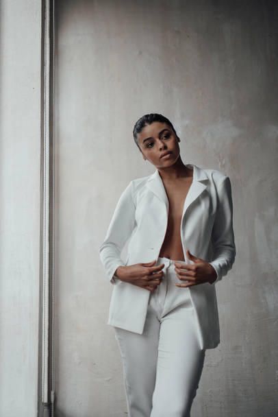 Young adult beauty swarthy woman in formal evening white suit with bra at thoughtful. Stylish black curly hair sensual african american model fashionista posing at studio in fashion pantsuit - Foto, Imagen