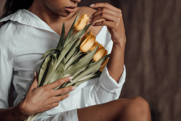 Mladá dospělá kráska snědá žena v bílé košili holá ramena objímají kytici žlutých tulipánů. Stylový černý kudrnaté vlasy smyslný africký americký model módní pózování ve studiu na jarní prázdniny - Fotografie, Obrázek