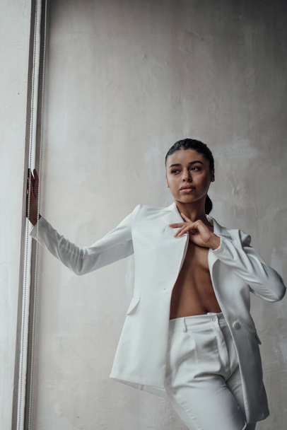 Young adult beauty swarthy woman in formal evening white suit with bra at thoughtful. Stylish black curly hair sensual african american model fashionista posing at studio in fashion pantsuit - Fotoğraf, Görsel