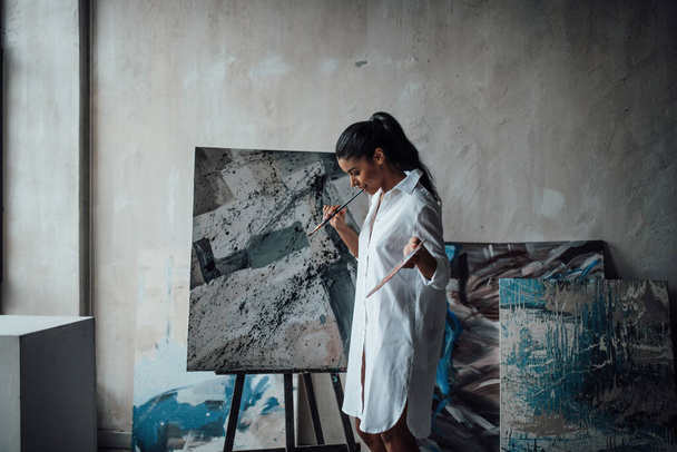 Fiatal művész szépség swarthy nő fehér hosszú ing csupasz lábak pózol neki festmények. Stílusos fekete göndör haj érzéki afro-amerikai modell festék paletta és ecsetek festés stúdió - Fotó, kép