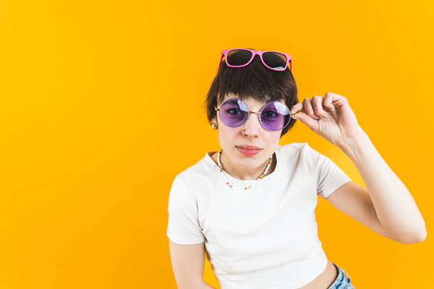 Femal model looking directly into the camera through a pair of sunglasses and wearing another pair on her head. Bright orange background. Medium shot. High quality photo - Photo, Image