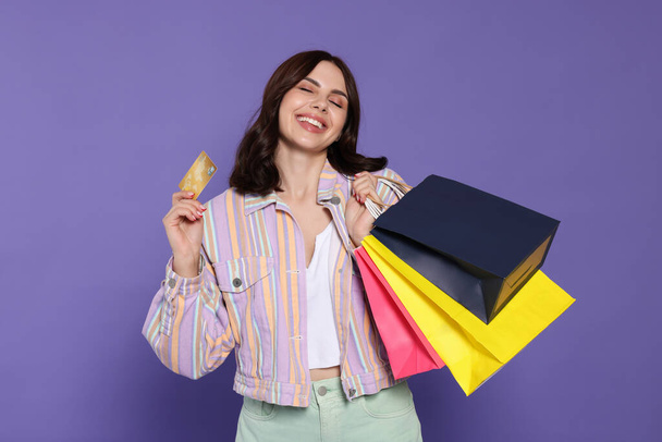 Beautiful young woman with paper shopping bags and credit card on purple background - Photo, image