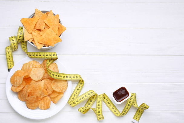 Potato chips, tomato sauce and measuring tape on white wooden table, flat lay with space for text. Weight loss concept - Фото, зображення