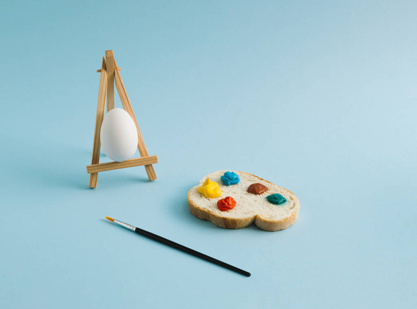 Abstrakcyjna kompozycja wielkanocna wykonana z białego jajka na sztaludze i kromki chleba z kolorową paletą. Pastelowe niebieskie tło. Przestrzeń kopiowania. - Zdjęcie, obraz