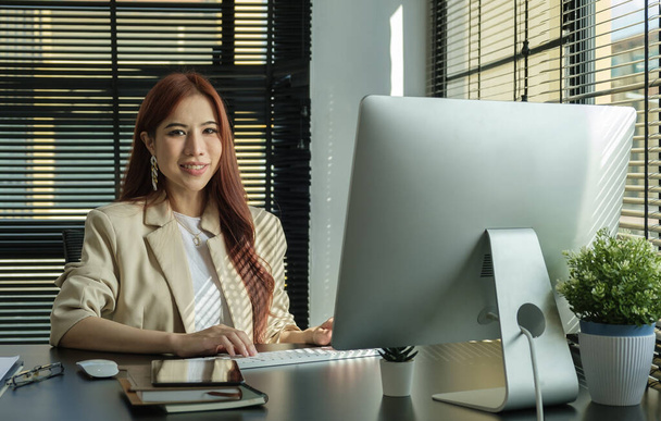 Επιτυχημένη Διευθύνων Σύμβουλος γυναίκα σε κομψό κοστούμι κάθεται στο σύγχρονο χώρο εργασίας και ψάχνει με σιγουριά στην κάμερα. - Φωτογραφία, εικόνα
