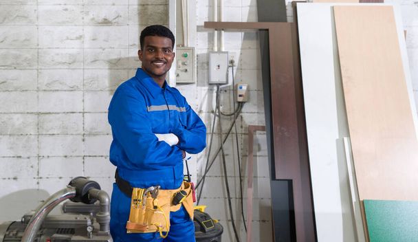 Young technician in blue mechanic jumpsuit, protective glove and yellow Tool Belts stand with arms crossed in a wooden furniture factory. - Foto, Bild
