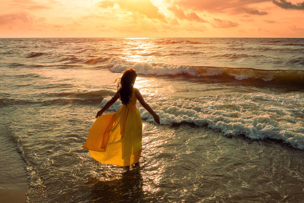 Seascape during golden sunrise with beautiful sky. Woman on the beach. Young happy woman in a yellow fluttering dress walks along the seashore. The girl looks at the magical sunrise. View from above. - Photo, image