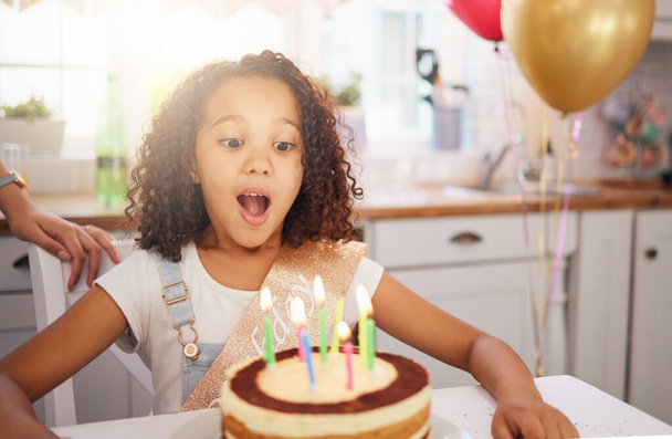 Happy birthday cake, candles and child with surprise, excited and happy face for love, care and celebration. Party, balloons and celebrate with black girl kid and dessert at the kitchen house table. - 写真・画像