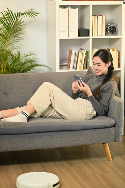 Ευτυχισμένη νεαρή γυναίκα χαλαρώνοντας στον καναπέ και κουβεντιάζοντας στο διαδίκτυο στο κινητό τηλέφωνο. 'Ανθρωποι, τεχνολογία και τρόπος ζωής. - Φωτογραφία, εικόνα