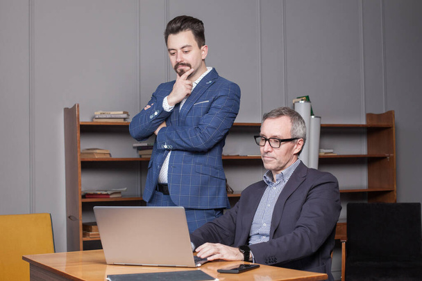 Dojrzały siwowłosy biznesmen w niebieskim garniturze i okularach do oczu ze swoim partnerem biznesowym są pensivly myślenia przez laptop w biurze - Zdjęcie, obraz