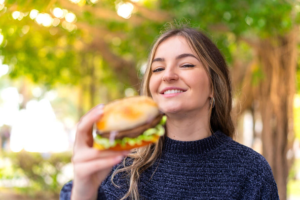 Joven mujer rumana bonita sosteniendo una hamburguesa al aire libre con expresión feliz - Foto, imagen