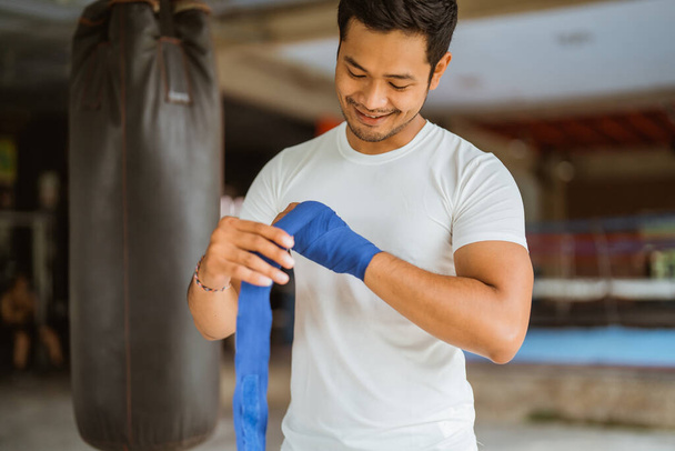 a asian male boxer wearing the boxing wrap gloves while standing with the box ring at the background - Foto, imagen