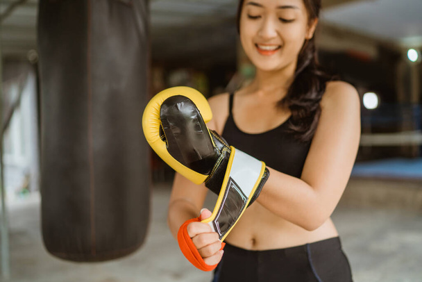 a female boxer standing with the hand wraps at her right hand while wearing the boxing gloves at her left hand inside the boxing studio - Photo, Image