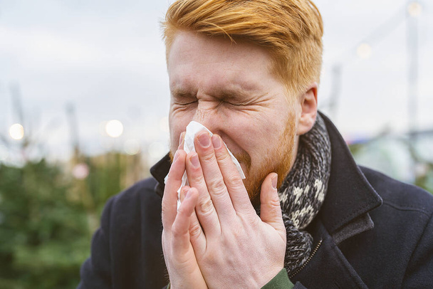 Ο άνθρωπος φτερνίζεται και φυσάει τη μύτη του σε ένα μαντήλι το χειμώνα με αλλεργία ή μόλυνση από ιό. Πορτρέτο της κοκκινομάλλα άνθρωπος εξωτερική. - Φωτογραφία, εικόνα