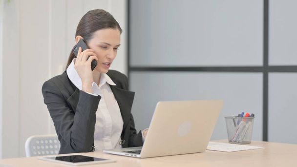Бізнес-леді розмовляє по телефону під час використання ноутбука
 - Фото, зображення