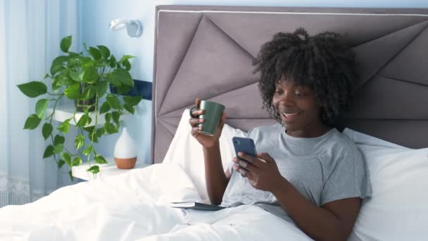 Usmívající se africká mladá žena dělá videohovor na smartphone a pití kávy v posteli v dopoledních hodinách. - Záběry, video