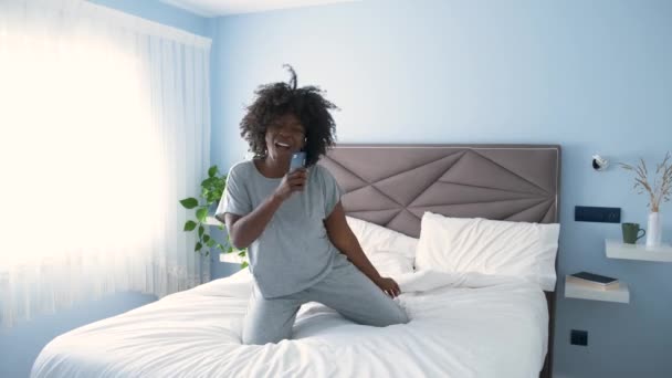 Mujer joven africana sonriente con pijama bailando con música favorita en la cama por la mañana. - Metraje, vídeo