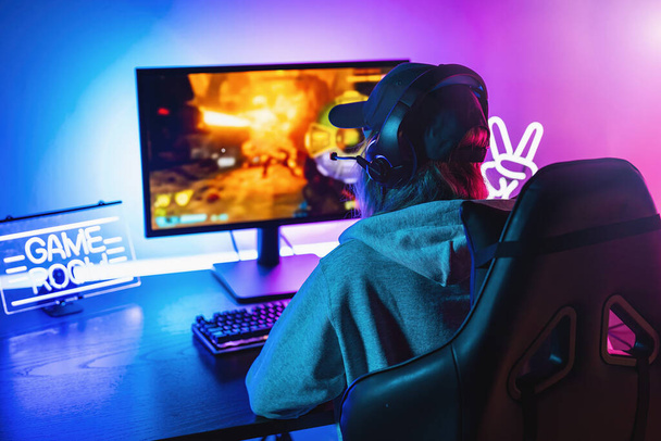 Chica jugando en primera persona Shooter Juego de vídeo en línea en su potente ordenador personal. La habitación y la PC tienen luces led de neón coloridas. Mujer joven está jugando con una gorra en casa - Foto, imagen