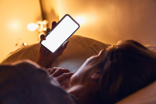 Mujer sosteniendo teléfono inteligente tarde en la noche con fondo blanco para copyspace en la cama. Persona mirando la aplicación de teléfono en blanco en el hogar oscuro. Imagen de concepto de sexting o acecho. - Foto, imagen