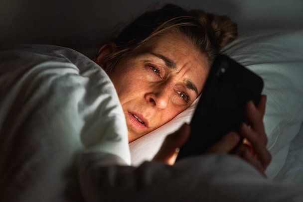 Nuori nainen käyttää älypuhelinta makatessaan sängyssä yöllä. Unettomuus ja sosiaalisen median riippuvuus käsite.  - Valokuva, kuva