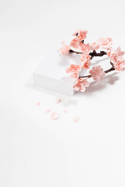 Spring stage mockup - square podium for presentation cosmetic products, goods, twig of tender pink sakura flowers, petals on white wood background in japanese traditional style, top view, vertical.  - Photo, Image