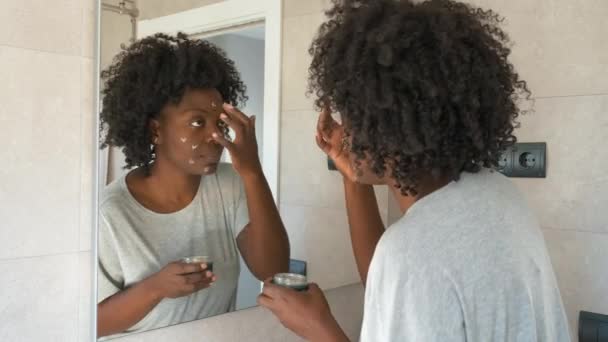 Heureuse jeune femme africaine appliquant une crème faciale écologique dans la salle de bain. - Séquence, vidéo