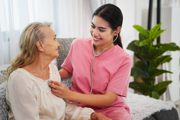 Elderly woman and nurse at home. Healthcare worker girl cares about senior female person indoors - Foto, immagini