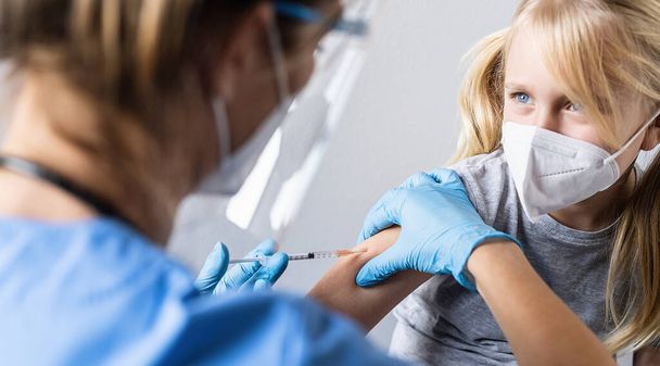 予防接種を受けている間、またはcovid-19パンデミックの間に撃たれている間、ブロンドの子供の女の子は手に注射器で医者を見て、 - 写真・画像