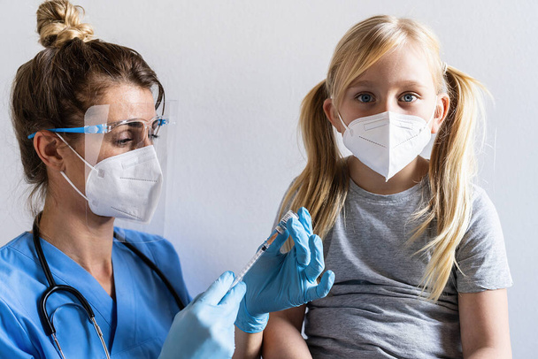 フェイスマスクをしたブロンドの女の子は、予防接種センターがワクチン接種を受けることに驚いています。covid-19コロナウイルス、インフルエンザ、危険な感染症のワクチンで注射する.   - 写真・画像