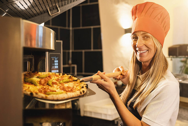 παραδοσιακή γυναικεία πίτσα αρτοποιός κρατώντας μια φρέσκια πίτσα από τον πέτρινο φούρνο με φλούδα στην ιταλική πιτσαρία - Φωτογραφία, εικόνα