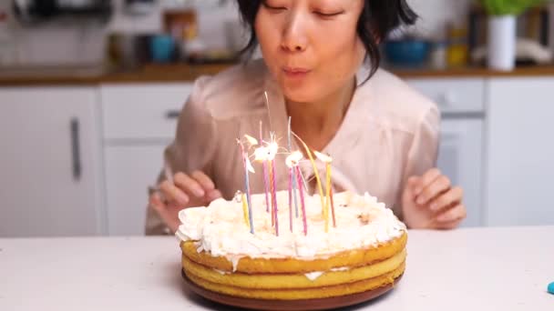 Primo piano della donna asiatica a occhi chiusi che esprime desideri e soffia candele sulla torta di compleanno a casa. Gioia e stile di vita. - Filmati, video