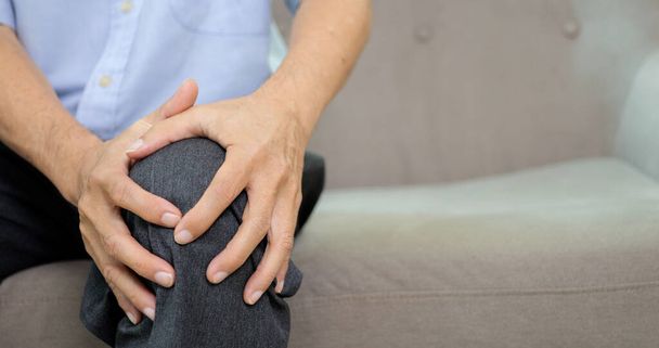 Zbliżenie ujęcia seniora cierpiącego na ból kolana siedzącego na kanapie w domu, Starszy mężczyzna dotykający kolana w punkcie bólu, Ludzie z koncepcją problemu zdrowotnego - Zdjęcie, obraz