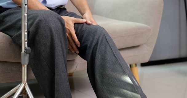 Старший дідусь, який ходив з палицею, намагаючись вправляти коліно і ногу на дивані вдома, літня людина з болем і стражданнями на колінах. - Фото, зображення