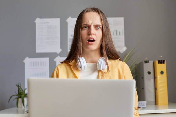 Image of surprised shocked scared woman wearing yellow shirt sitting in office in front of her notebook, looking at camera with shocked face and open mouth, having problems with her work. - Фото, зображення