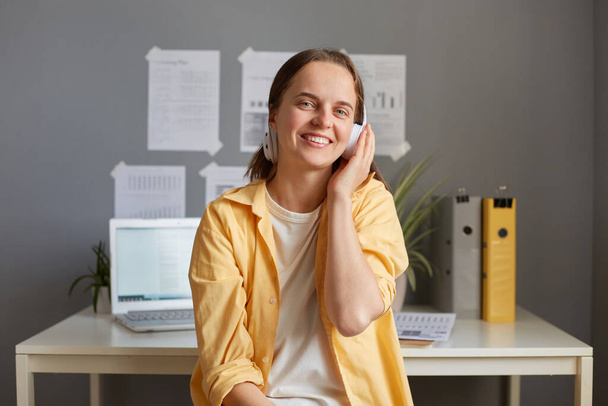 Portrait of beautiful female freelancer sitting in office while having break, listening to music or podcast, wearing casual clothing, looking at camera, holding headphones. - Foto, Bild