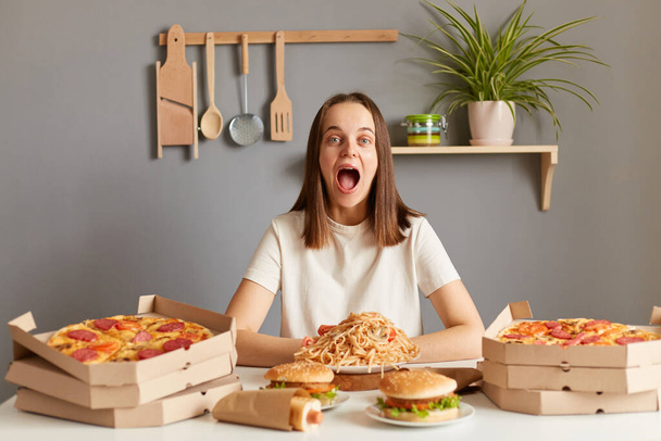 Indoor shot of amazed excited extremely happy Caucasian woman wearing white casual T-shirt sitting at table in kitchen, breaking diet, having lots takeaway fast food. - 写真・画像