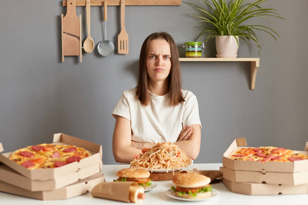 Portrait of sad offended Caucasian woman wearing white casual T-shirt sitting at table with big variety of fast food, keeps arms folded, can't eating, keeps diet, looking at camera with sorrow. - Valokuva, kuva