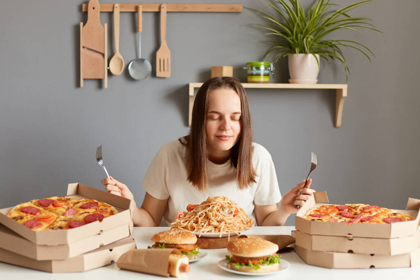 Horizontal shot of satisfied woman with brown hair wearing white casual T-shirt sitting at table in kitchen, posing with closed eyes, smelling pasta, pizza, hamburgers. - 写真・画像